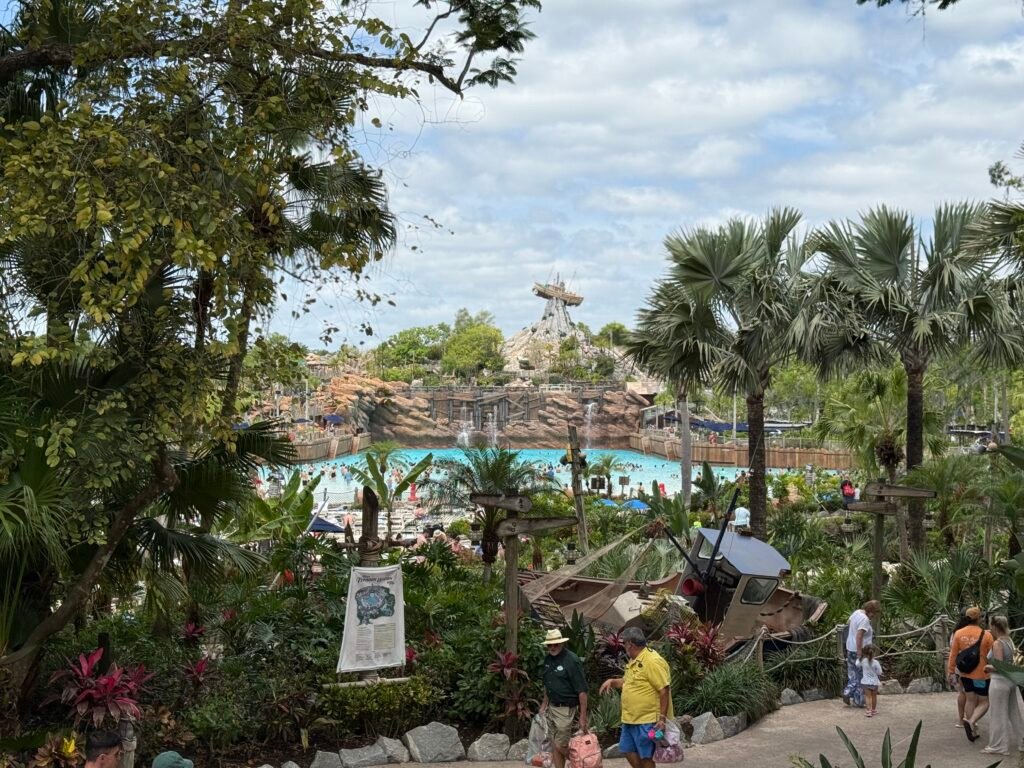 View of Typhoon Lagoon Water Park with lush greenery, a sandy area, and a pool visible in the background. Some guests are enjoying the park.